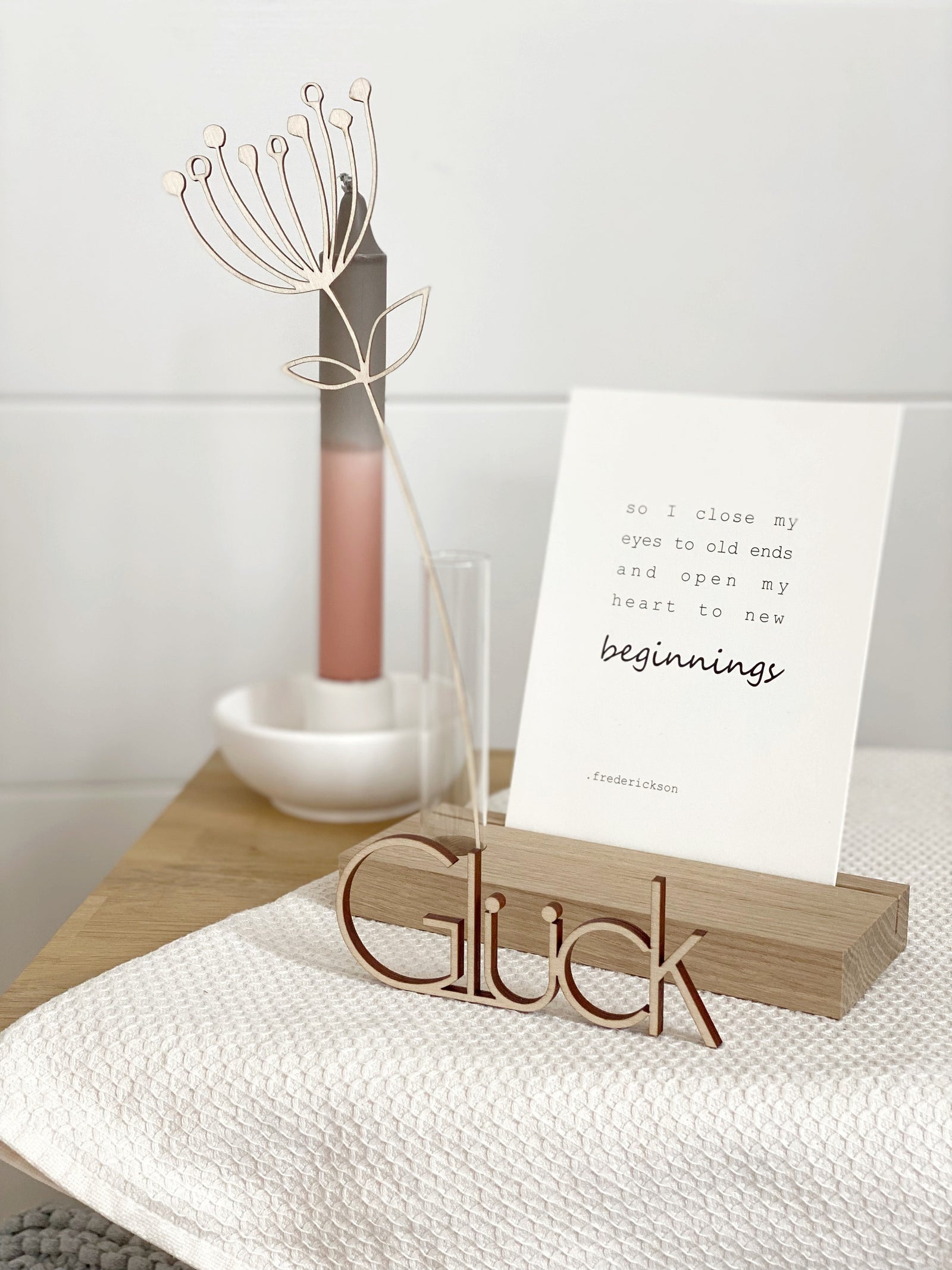 Inspiration : Kartenhalter aus Eiche mit Postkarte und einem Reagenzglas mit gelaserter Holzblume. Davor ein Schriftzug "Glück" aus Holz, dahinter eine weiße Kerzenschale mit zweifarbiger Kerze