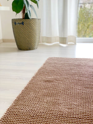 Leinenfarbener Teppich aus groben Recyclinggarn auf einem weißen Boden. Im Hintergrund eine Pfanze in einem Bastkorb. Beispiel für eine  individuelle Anfertigung von Stadtrandstil  handmade