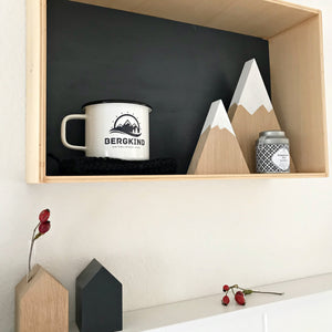 Dekorationsinspiration: ein Regal, bestückt mit zwei unterschiedlich großen, stilisierten Bergen aus Eichenholz, einer Teedose und einer Emailletasse mit dem Schriftzug Bergkind. Darunter auf einem Bord zwei Holzhäuser in schwarz und natur sowie getrockneten Hagebutten