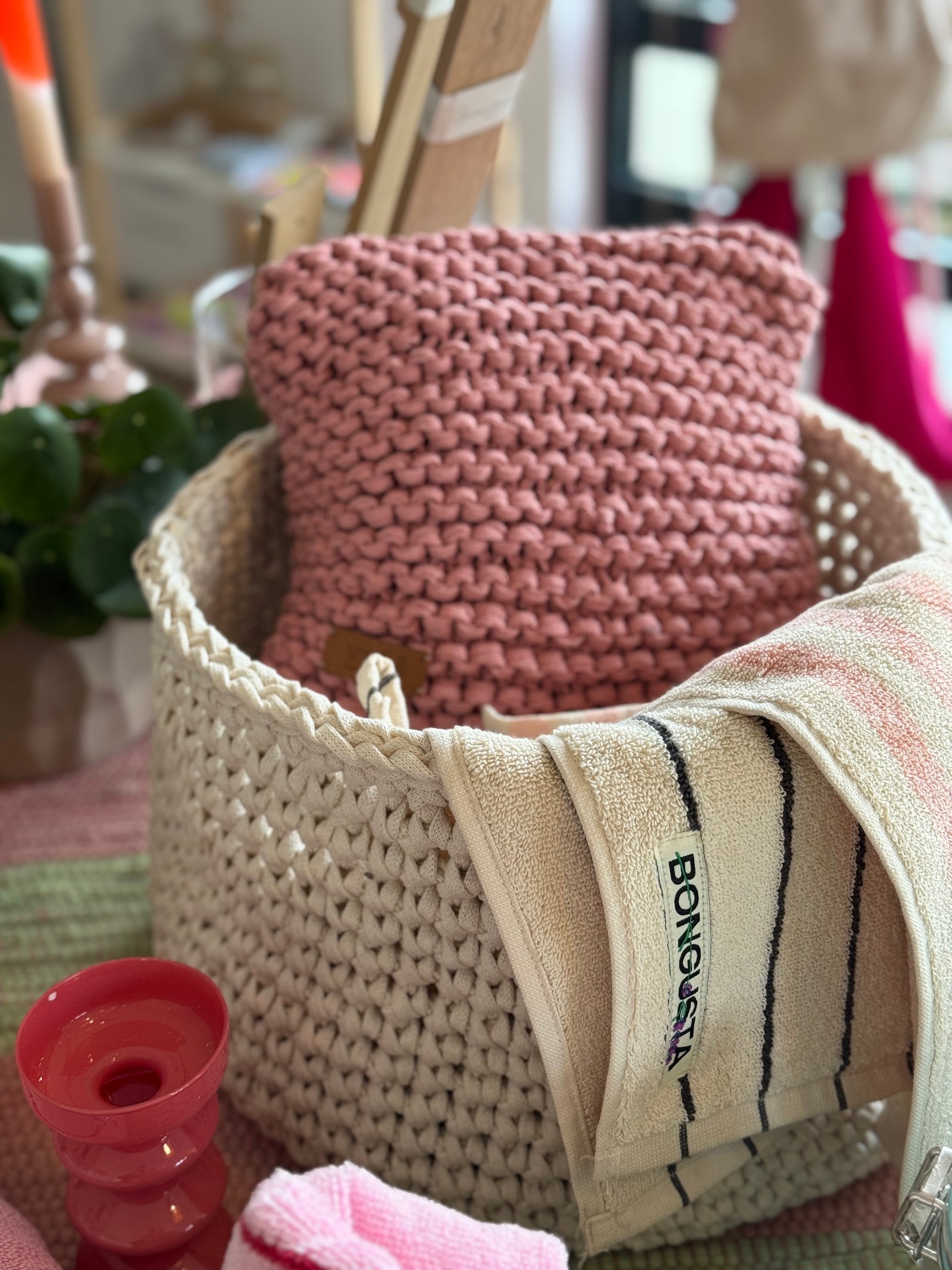 Naturfarbener gehäkelter Korb gefüllt mit einem himbeerfarbenem gestrickten Kissen und einem gestreifte Handuch