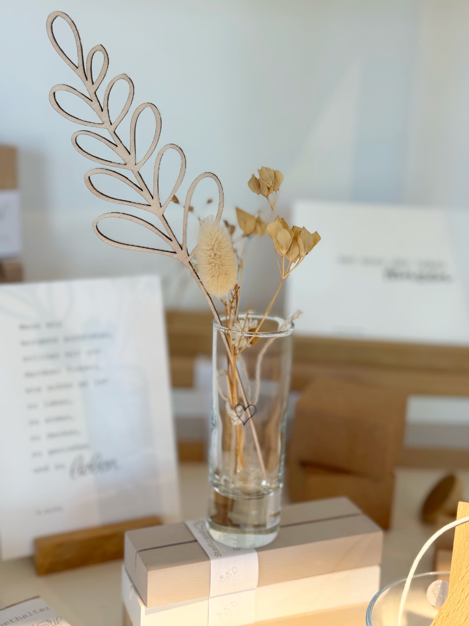 Schnapsglas mit Herz, das als Vase genutzt ist. Gefüllt mit Trockenblumen und einer Holzblume