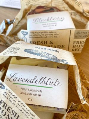 Zwei Seifenstücke in Papierverpackun, einmal Lavendelblüte, einmal Blackberry