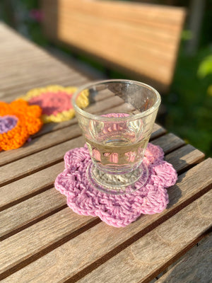 Stimmungsfoto : auf einem hölzernen Gartenisch liegt ein rosafarbener gehäkelter Untersetzer (handmade), darauf ein Glas. Im intergrund schemenhaft weitere Untersetzer in Sommerfarben
