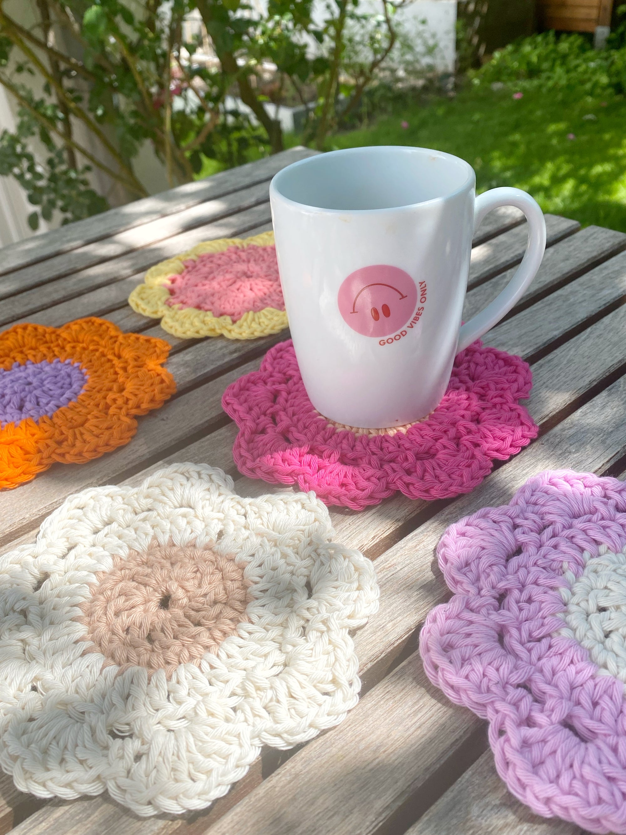 Gartentisch voller bunter Untersetzer in Blumenform, gehäkelt von Stadtrandstil handmade. Auf einem steht eine Tasse mit einem Smiley