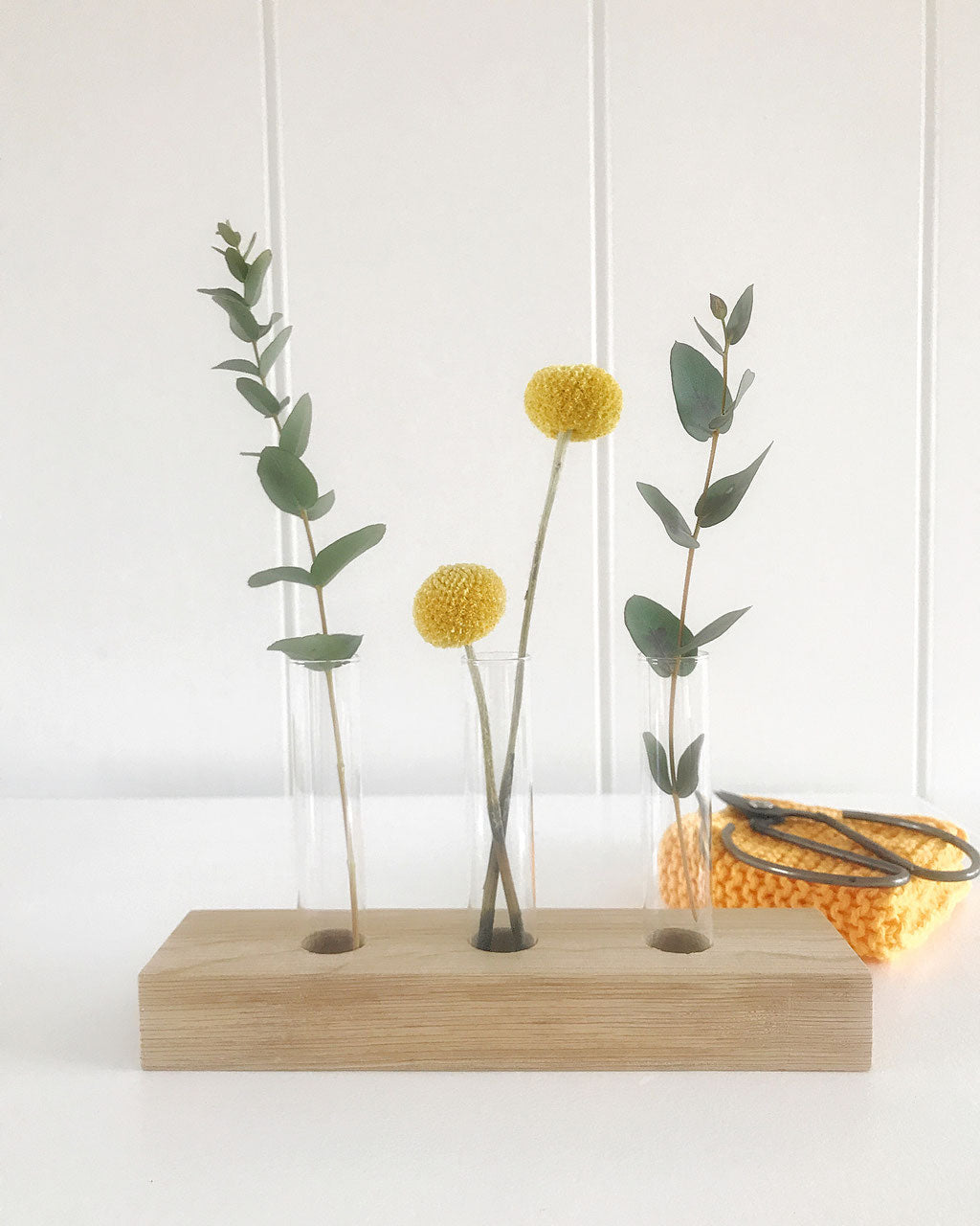 Blumenwiese; Blumendisplay mit drei Gläsern gefüllt mit Eukalyptus und Trommelstöcken, daneben ein kleines, gelbes Stricktuch und eine Schere