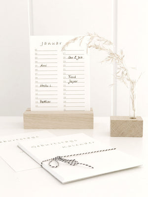 Geburtstagskalender (immerwährend) in minimalistischem Design von Stadtrandstil in Postkartenformat mit einem Halter aus unbehandeltem Eichenholz. Daneben eine Minivase mit getrocknetem Gras.