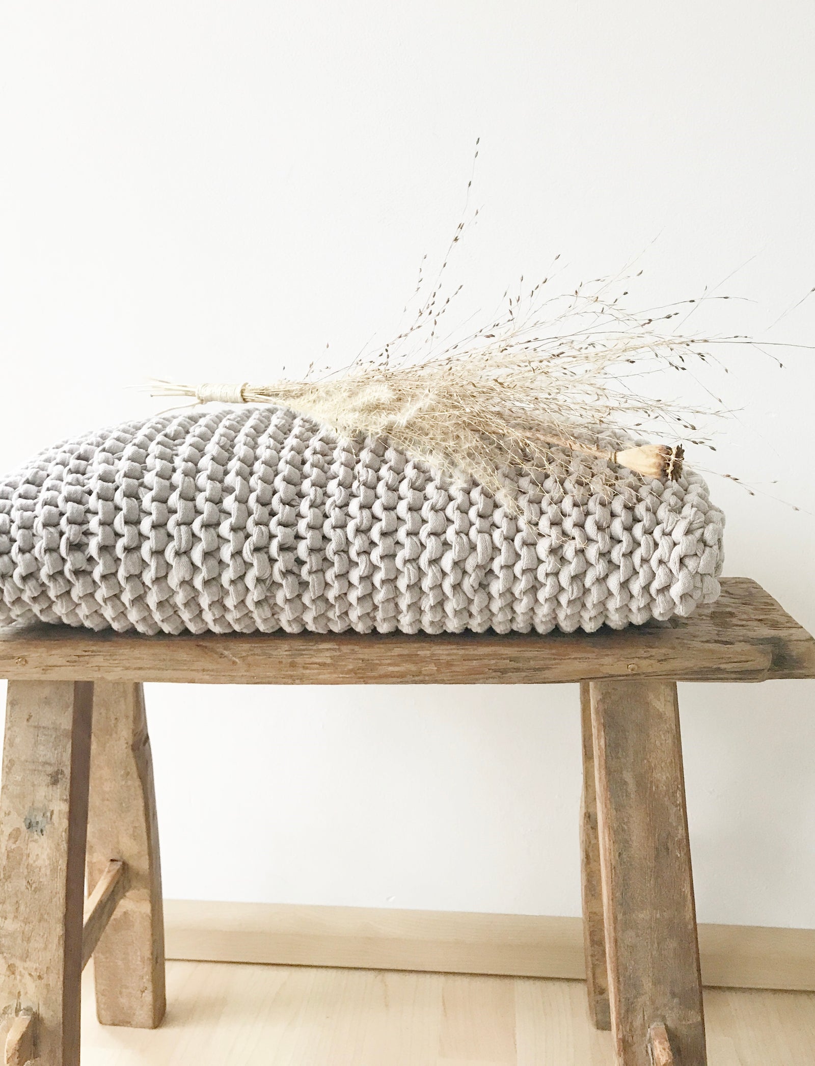 Naturfarbenes Kissen grob gestrickt auf einer Holzbank. Davor eine bauchige Glasvase mit getrockneten Gräsern. Eine natürliche Dekoration von Stadtrandstil