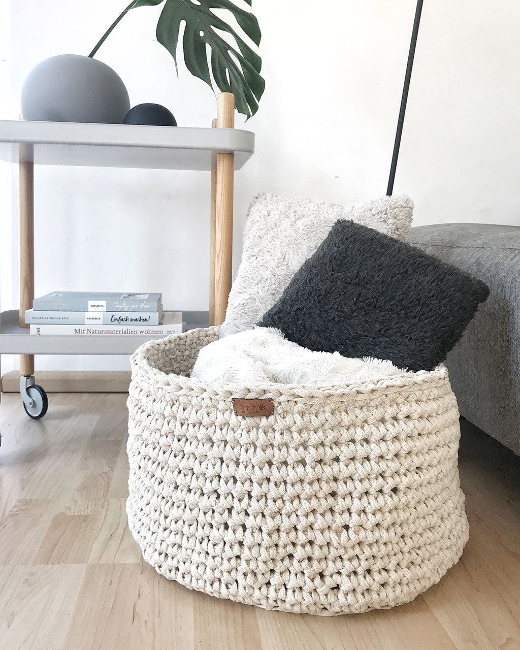 Wohnzimmer Inspiration im klaren Design: Moderner Beistellwagen mit Pflanze und Büchern dekoriert, eine schwarze Leselampe und dem extragroßem Häkelkorb von Stadtrandstil, in dem Kissen und eine Decke aufbewahrt werden