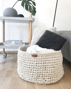 Natürlich, schlichtes Wohndesign mit einem großen, handgearbeitetem Aufbewahrungskorb mit Decken und Kissen. Dahinter ein Beistellwagen dekoriert mit Kugelvasen und Büchern