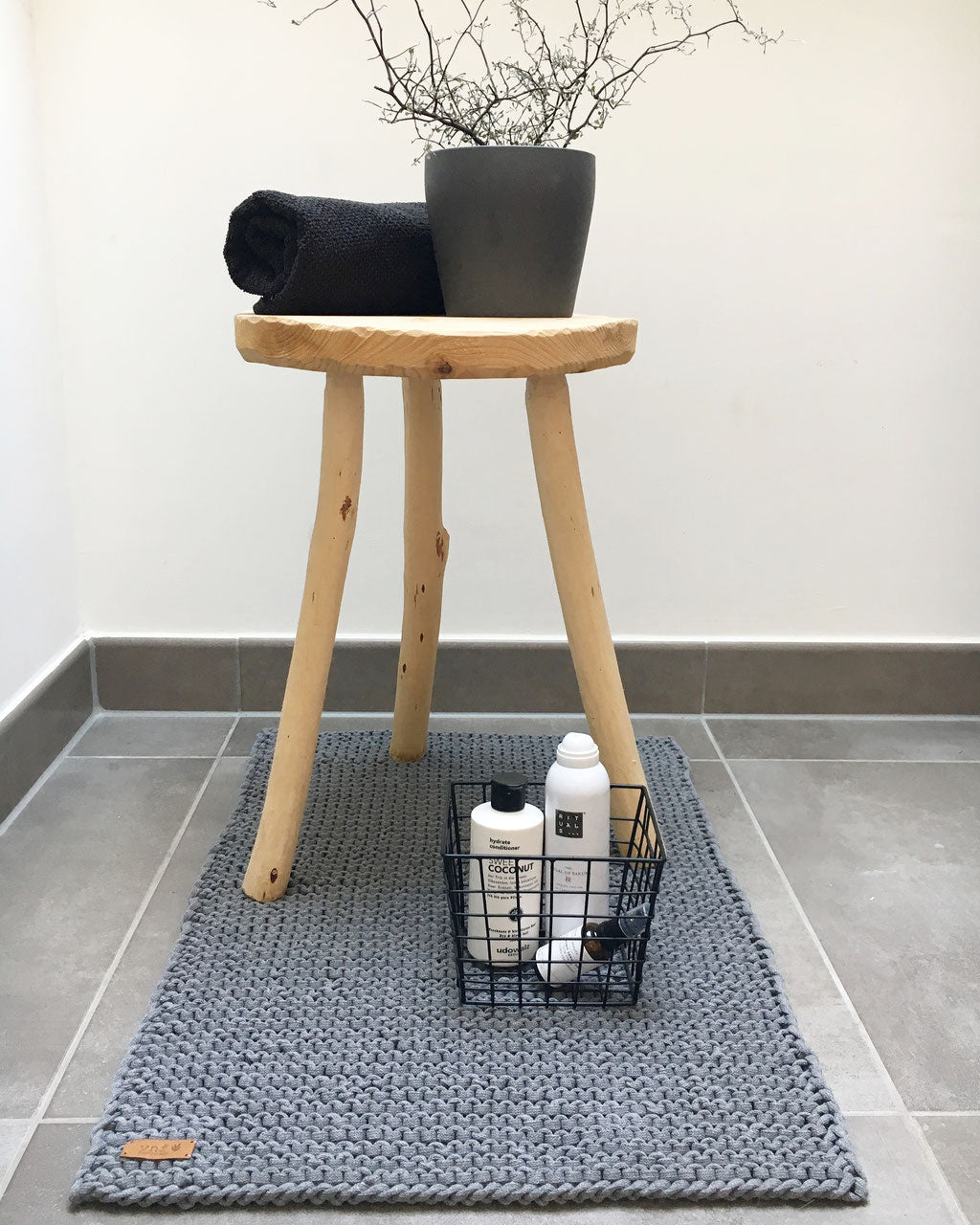 Dunkelgrauer Badvorleger aus Recyclinggarn.  Dekoriert mit Badezimmer Utensilien : einem Drahtkorb mit Pfelgeprodukten und einem Holzhocker mit schwarzem Badezimmer natürlich.andtuch und einer Pflanze. Badinspiraion Stadtrandstil
