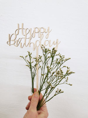 In der Hand wird ein kleiner Strauß mit weien Böüten gehalten sowie ien Caketopper aus gelasertem Holz "Happy Birthday"