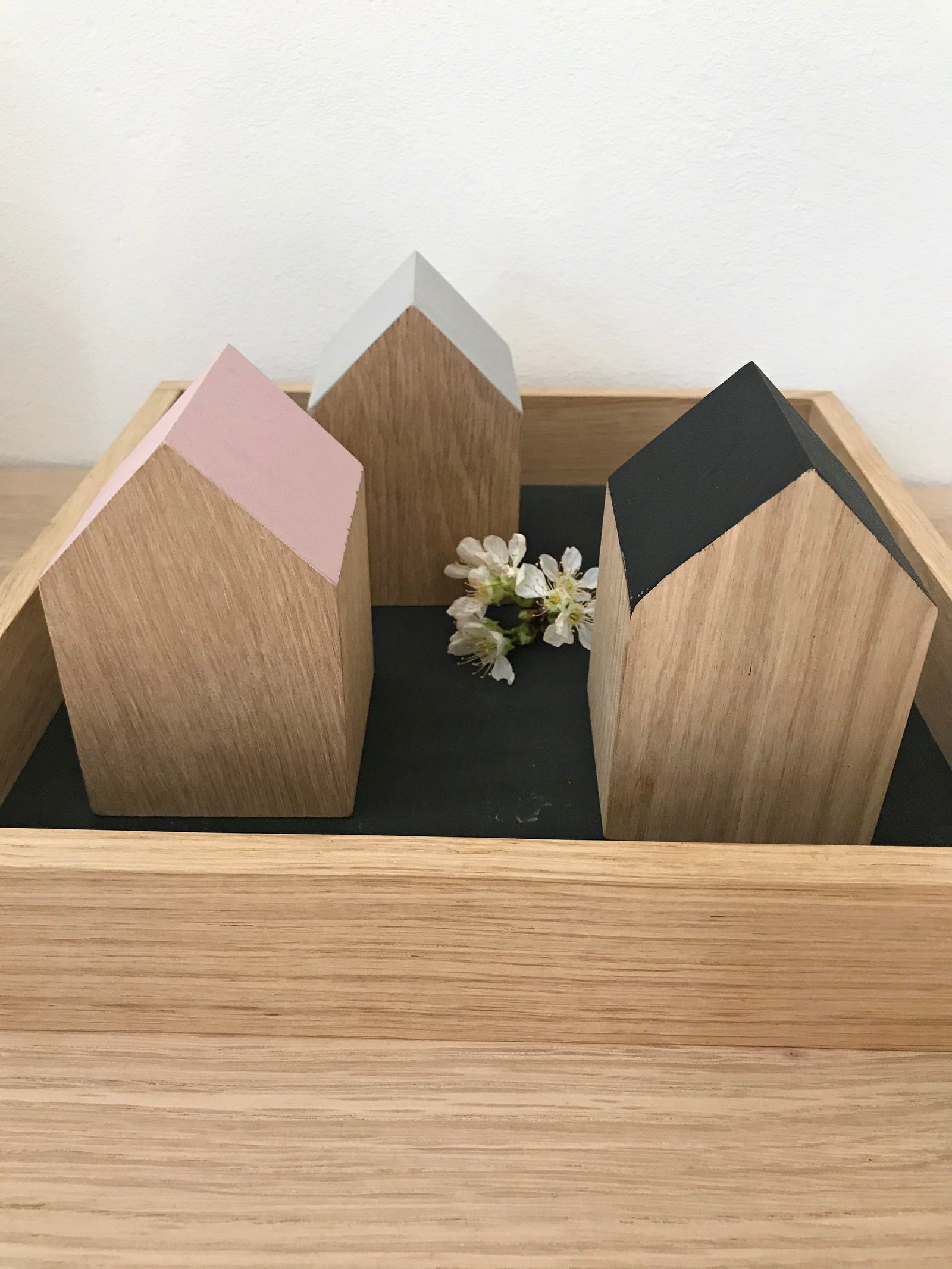 Drei kleine Dekohäuser aus Eichenholz, deren Giebel unterschiedlich lackiert sind. Je einmal in schwarz, rosé und sandstone. Sie stehen auf einem quadratischem Holztabeltt und sind mit einer zarten, kleinen Blume dekoiert