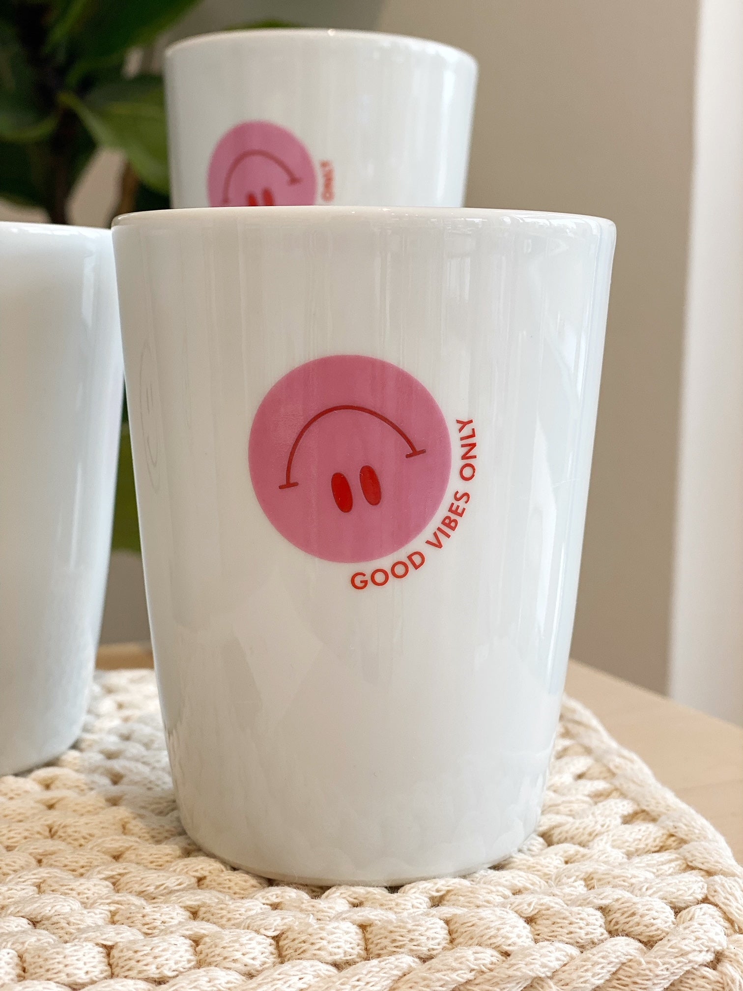 Fröhliches Arrangement mit weißen Bechern, auf die unterschiedliche Smileys gedruckt sind. Auf weißem Untersetzer, Grünpflanze im Hintergrund