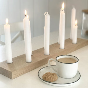 Stimmungsvolle Ansicht des großen Kerzenbretts von Stadtrandstil mit neun weißen, brennenden Kerzen. Davor eine weiße Tasse (gefüllt mit Kaffee) und Untertasse mit schwarzem Rand auf dem ein Keks liegt