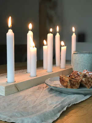 Dekoration Inspiration : gemütliche Atmosphäre mit Kerzenbrett aus Eiche und brennenden weißen Kerzen. Davor ein Kaffeebecher und ein Teller mit Kuchen auf einer naturfarbenen Leinendecke