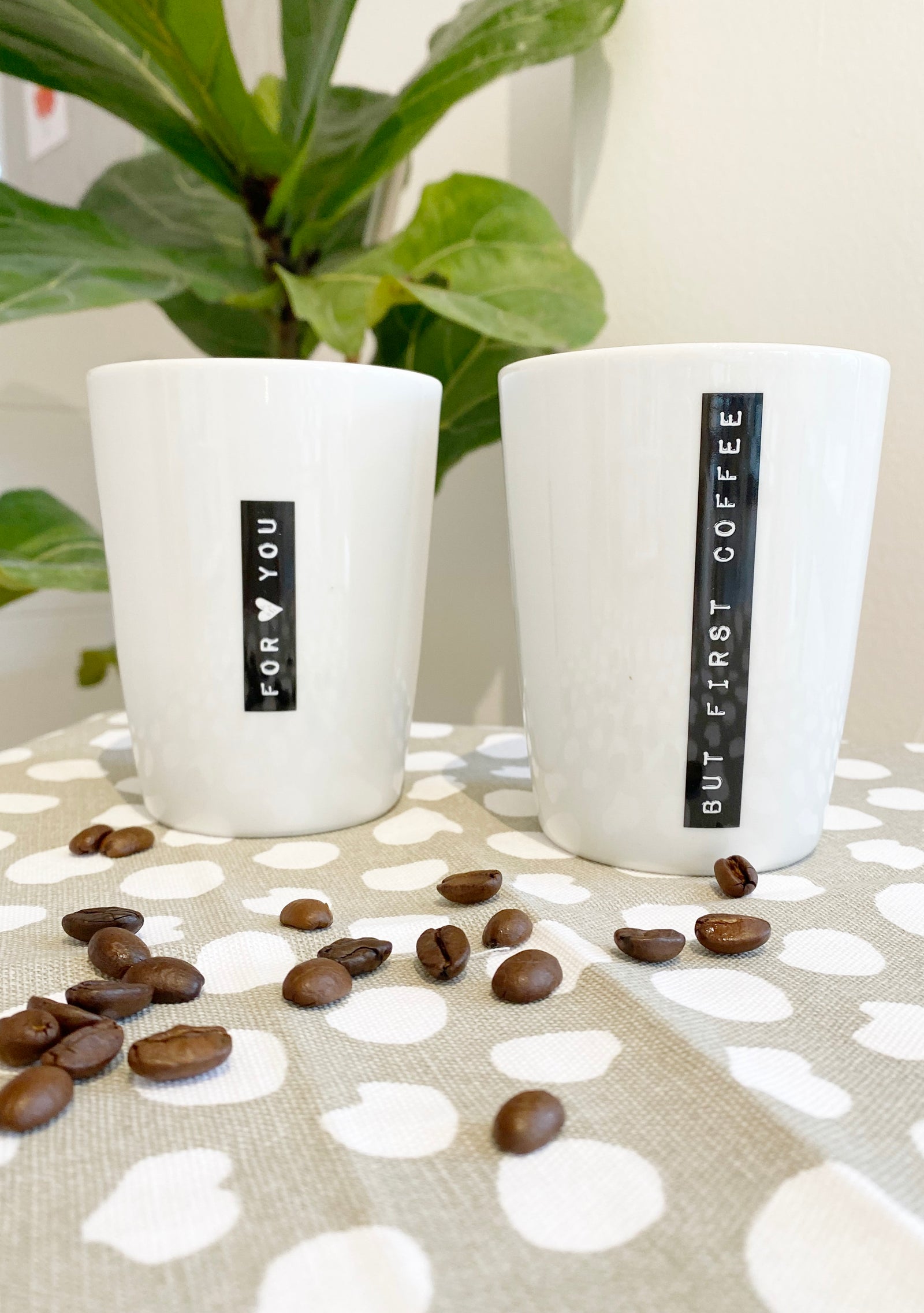 Zwei natürlich dekorierte Kaffeebecher mit dem schwarzen Aufdruck " for you" und "but first coffee", mit beige weißem Tuch, Kaffeebohnen und Grünpflanze