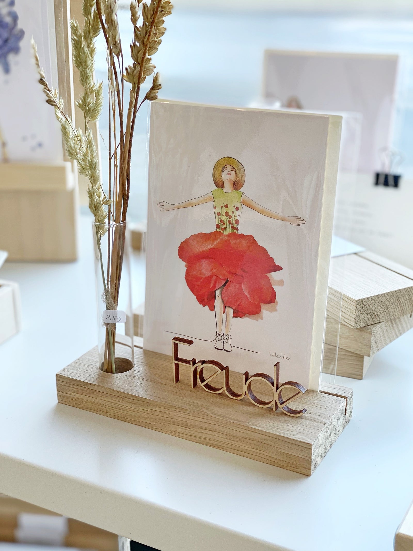 Inspiration : Kartenhalter aus Eiche mit Postkarte und einem Reagenzglas mit gelaserter Holzblume. Davor ein Schriftzug "Glück" aus Holz, dahinter eine weiße Kerzenschale mit zweifarbiger Kerze