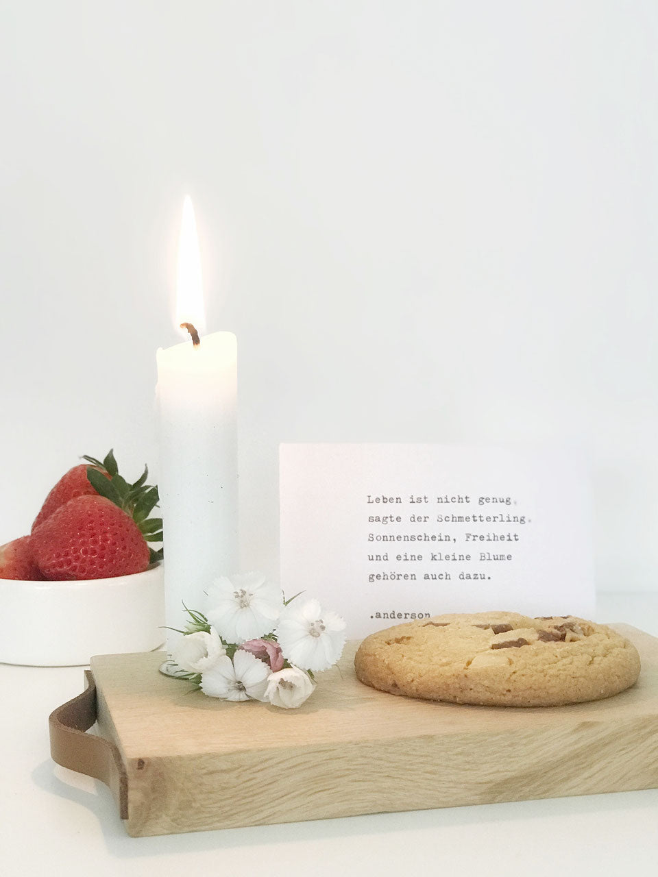 Kleines Kerzentablett mit einer weißen, brennenden Kerze auf dem ein paar bBlüten iegen und ein Keks. Dehinter eine Karte mit einem Zitat und ein paar Erdbeeren