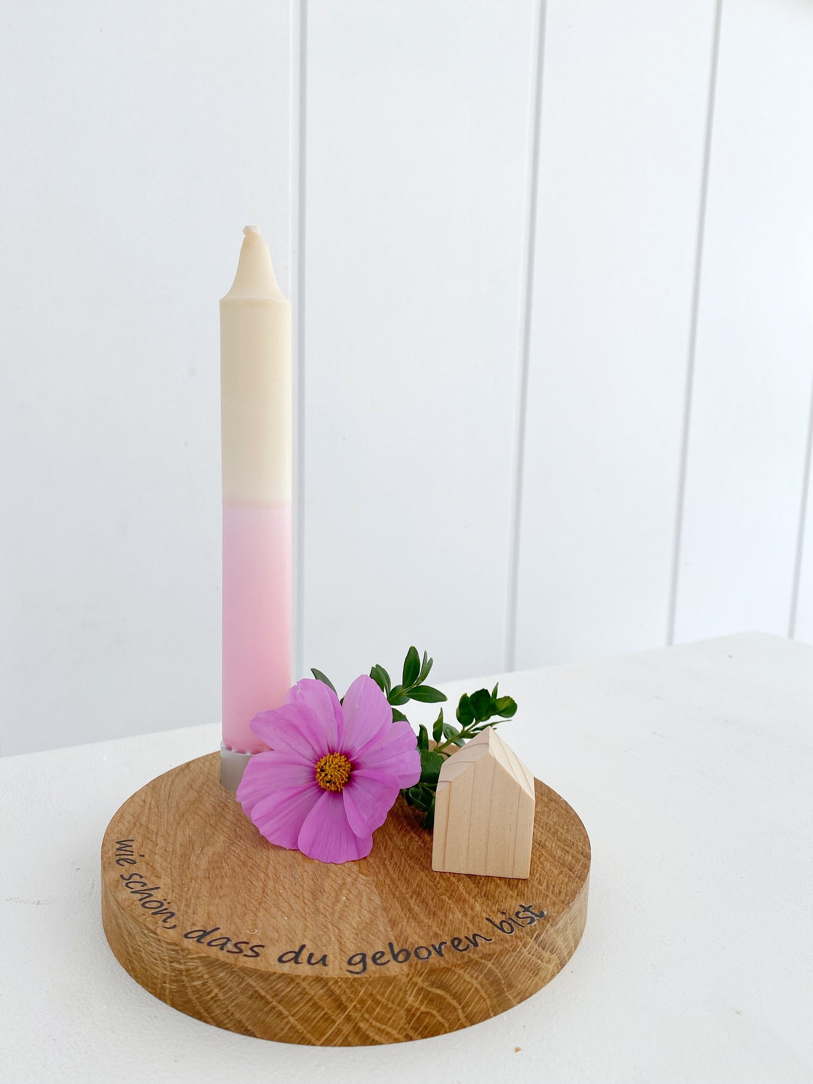 Rundes Brett aus Buche mit einem integrierten Kerzenhalter in dem eine pastellfarbene steht. Am Rand steht ein Schriftzug "wie schön, dass du geboren bist" (schwarze Plotterfolie), dekoriert mit einer rosafarbenen Kosmeeblüte und einem naturfarbenen Minihaus