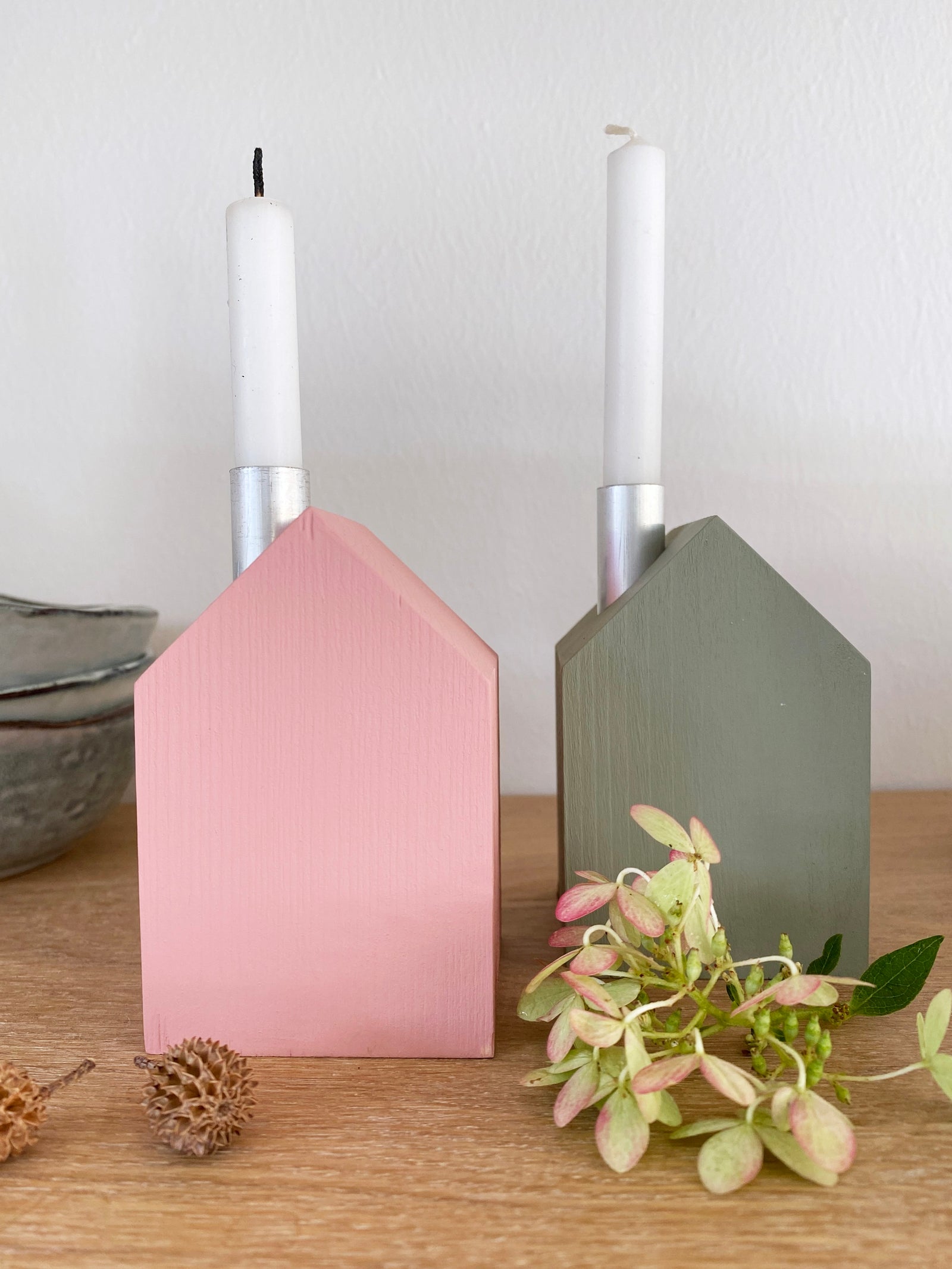 Zwei bunte Kerzenhäuser mit weißen Kerzen, eines in rosé, eines in oliv. Davor eine getrocknete Hortensienblüte