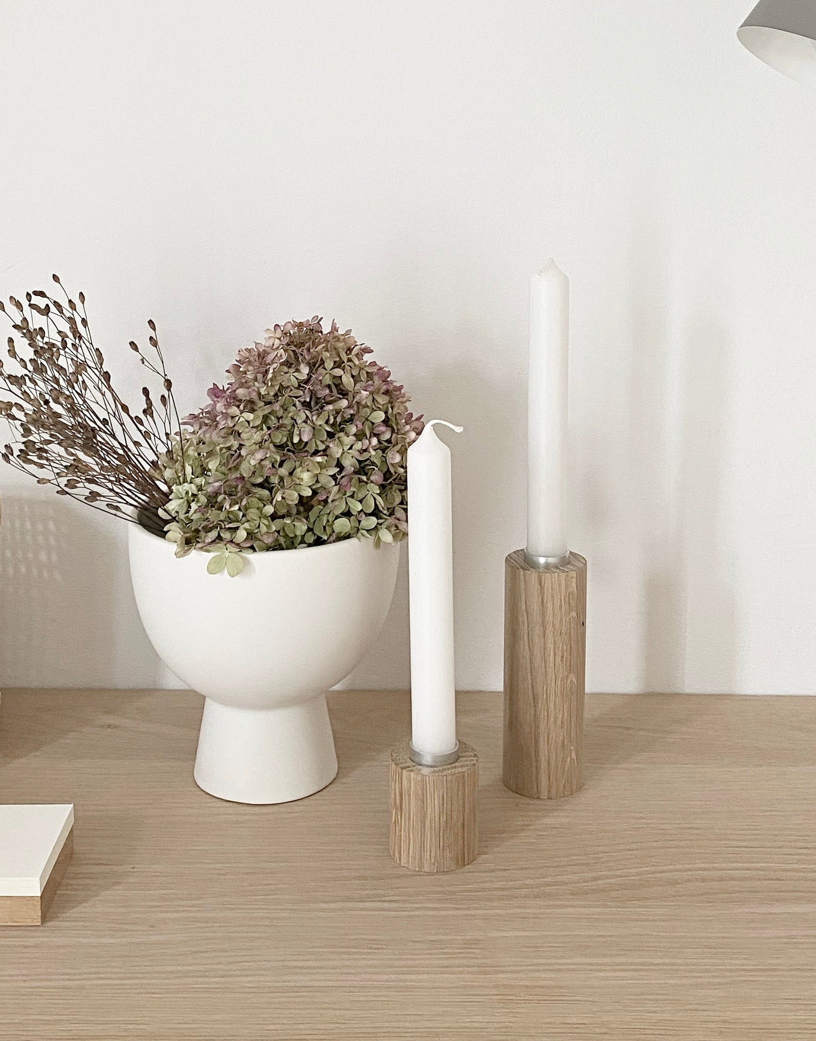 Zwei runde Kerzenständer aus Eiche in zwei Größen und mit weißen Kerzen stehen auf einem Eichentisch. Daneben eine weiße Schale auf Fuß, gefüllt mit einer getrockneten Hortensienblüte