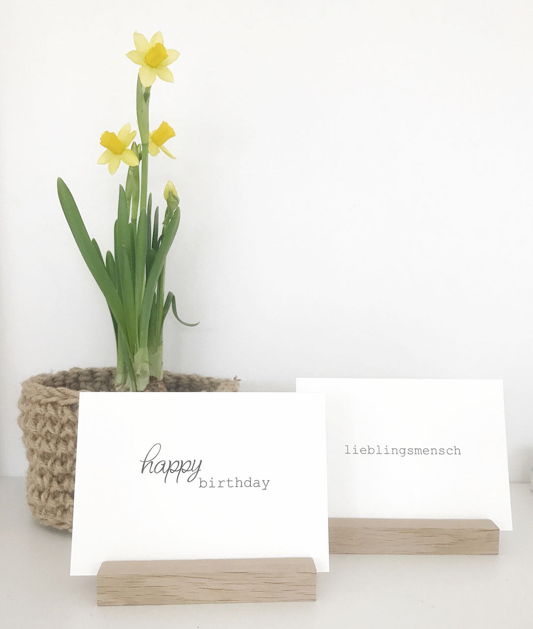 Minimalistsiche Fotohalter aus Eiche von Stadtrandstil mit je einer Postkarte. Dahinter ein Jutekorb mit Narzisse