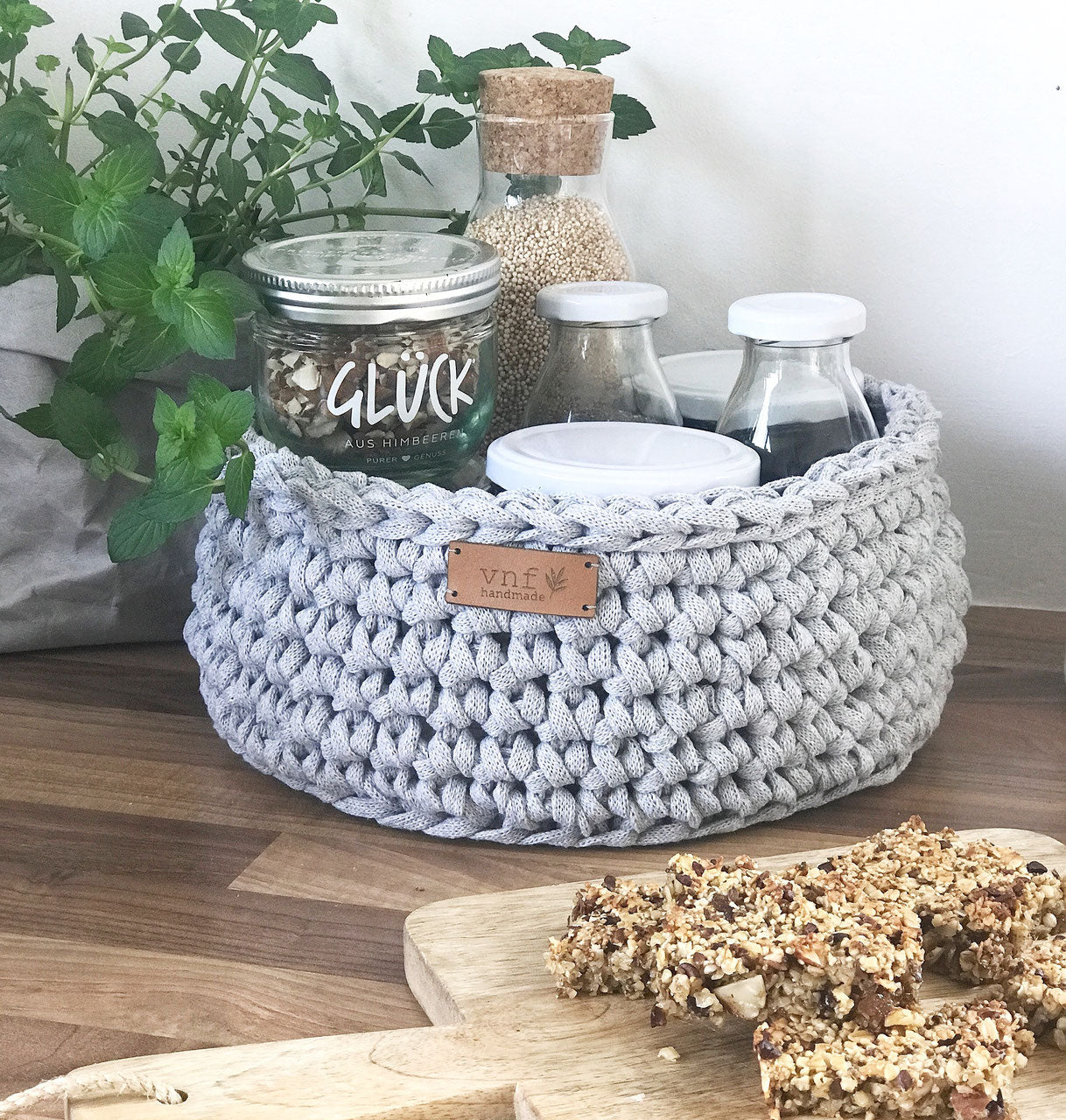 Hellgrauer Häkelkorb gefüllt mit Vorratsgläsern. Kücheninspiration mit Minzpflanze und einem Hlzbrett mit Gebäck