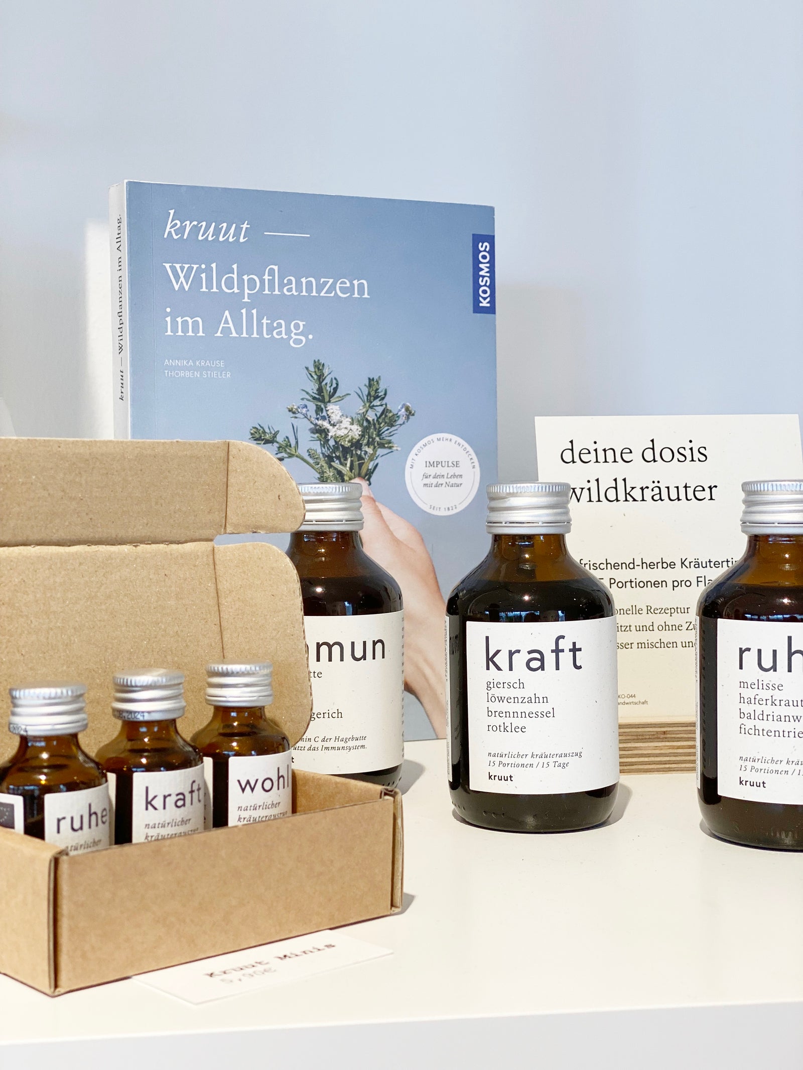 Setting mit mehreren Flaschen der Oxymels von Kruut. Dahinter das Buch kruut- Wildpflanzen im Alltag und ein Infozettel