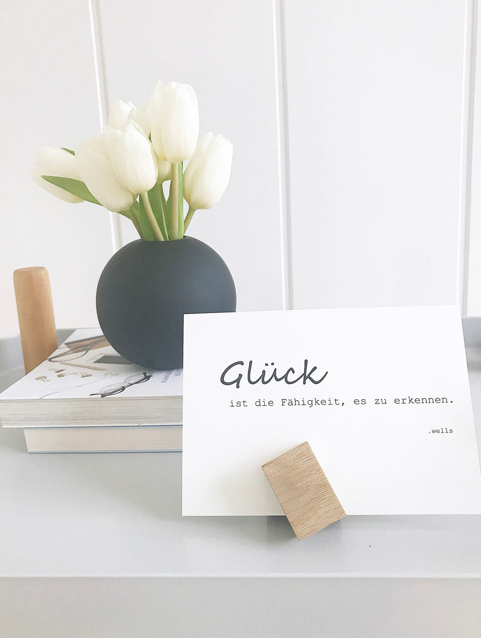 Zwei Magnethalter aus Eiche halten eine Postkarte. Dahinter eine schwarze Kugelvase mit weißen Tulpen und ein paar Bücher