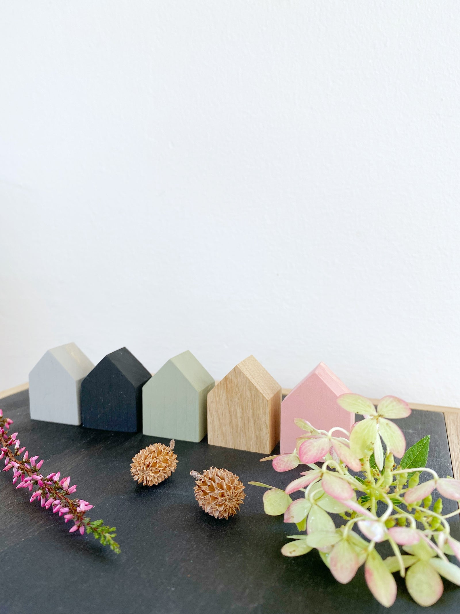Fünf Miniäuser in unterschiedlichen Farben auf schwarzem Untergrund. Dekoriert mit rosafarbenen Pflanzen und kleinen Zapfen