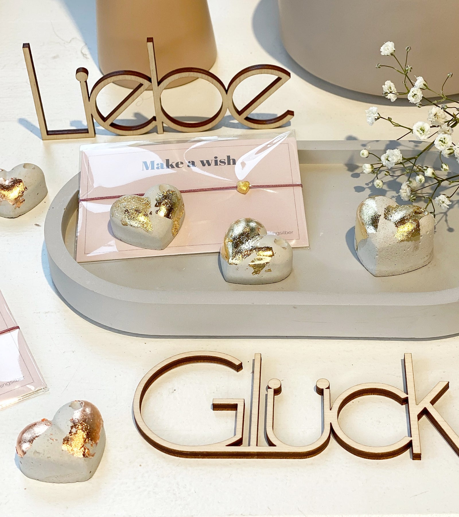 Hübsch dekoriert: ein ovales Beton tablett mt drei Betonherzen mit Blattgold, einem Armband und Schleierkraut. Dhinter steht ein Schriftzug "Liebe" aus Holz, davor ein Schriftzug, liegend "Glück"