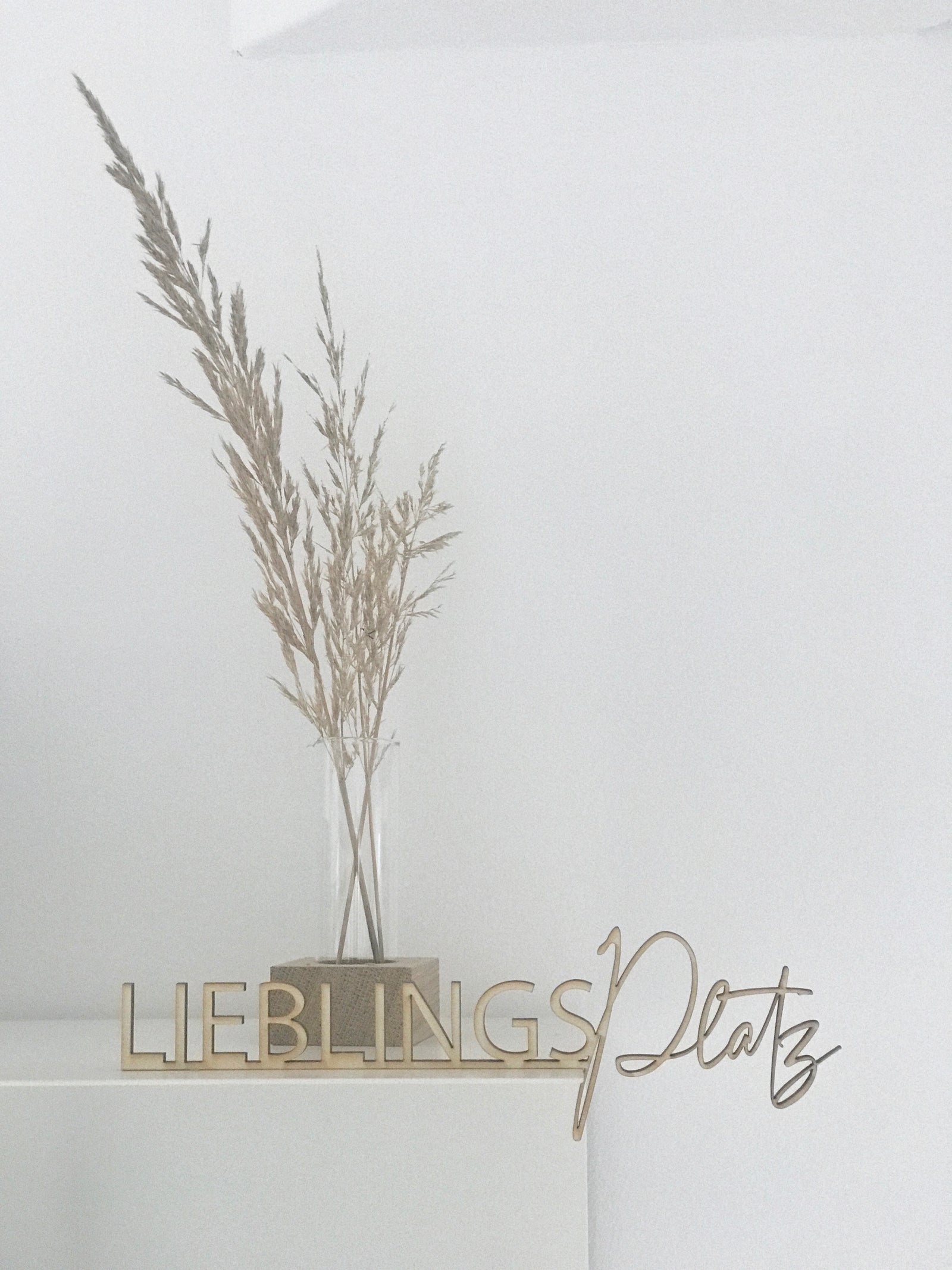 zweifarbige Dekoration mit einem Holzschriftzug "lieblingsplatz" und einer kleinen Holvase, gefüllt mit getrockneten Gräsern