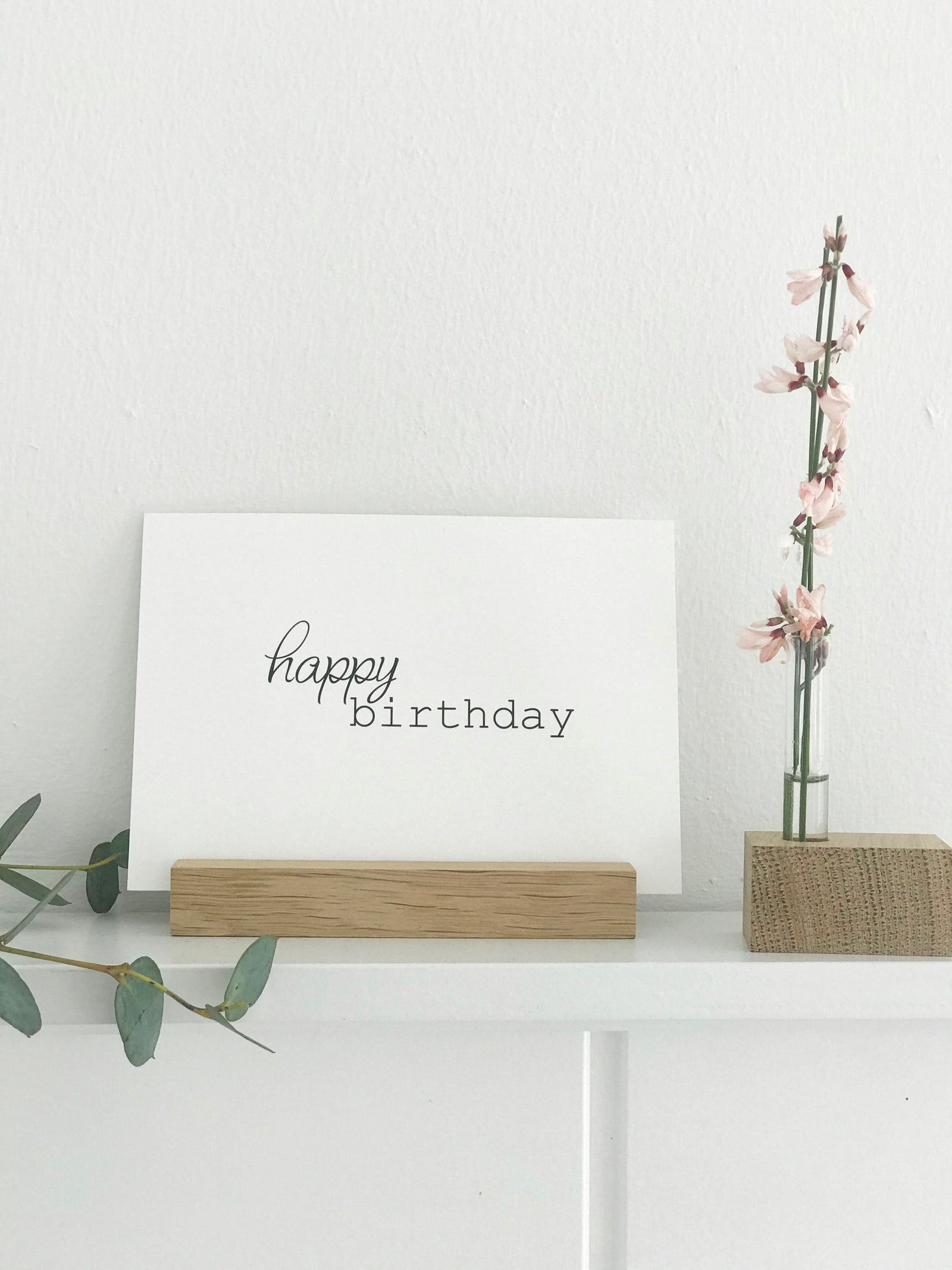 auf einem weißen Bord steht ein Kartenhalter aus Eiche von Stadtrandstil handmade mit einer Postkarte mit dem Text "happy birthday", daneben eine Minivase mit einem Zweig rosa Ginster