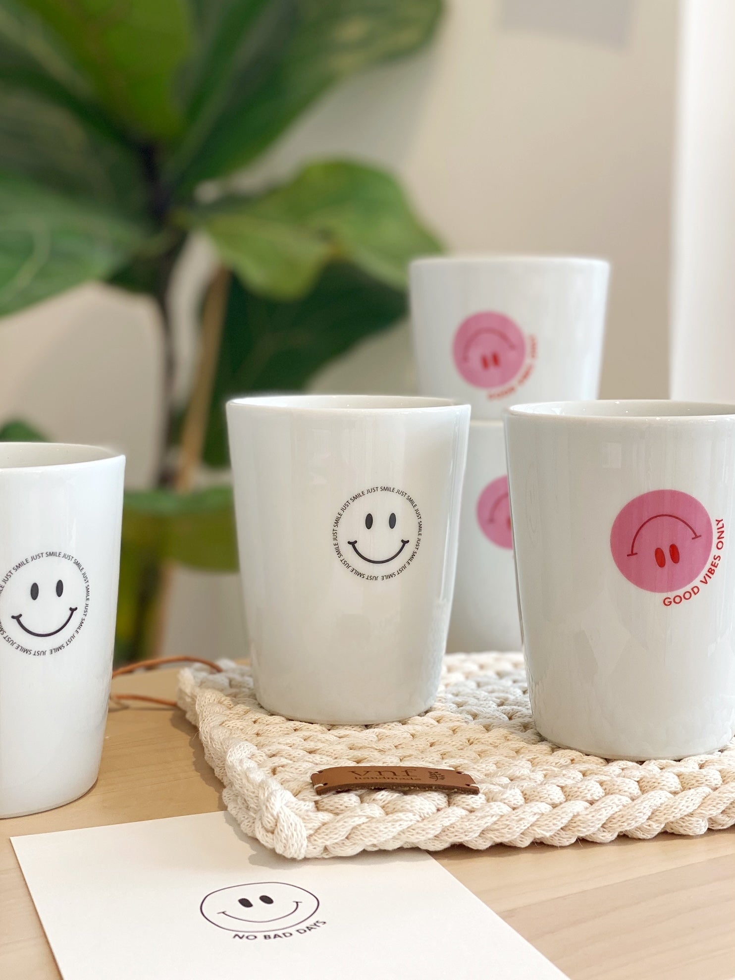 Fröhliches Arrangement mit weißen Bechern, auf die unterschiedliche Smileys gedruckt sind. Auf weißem Untersetzer, Grünpflanze im Hintergrund