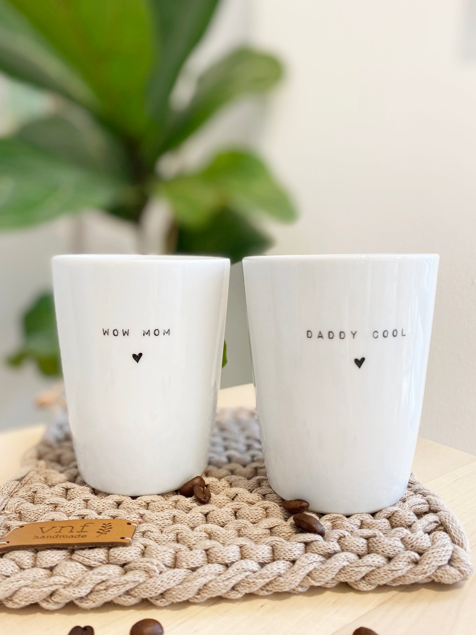 zwei weoße Becher aus Porzellan mit dem Aufdruck "wow mom" und "daddy cool". Natürlich arrangiert auf handgestrickten Untersetzer mit Grünpflanze und Kaffeebohnen