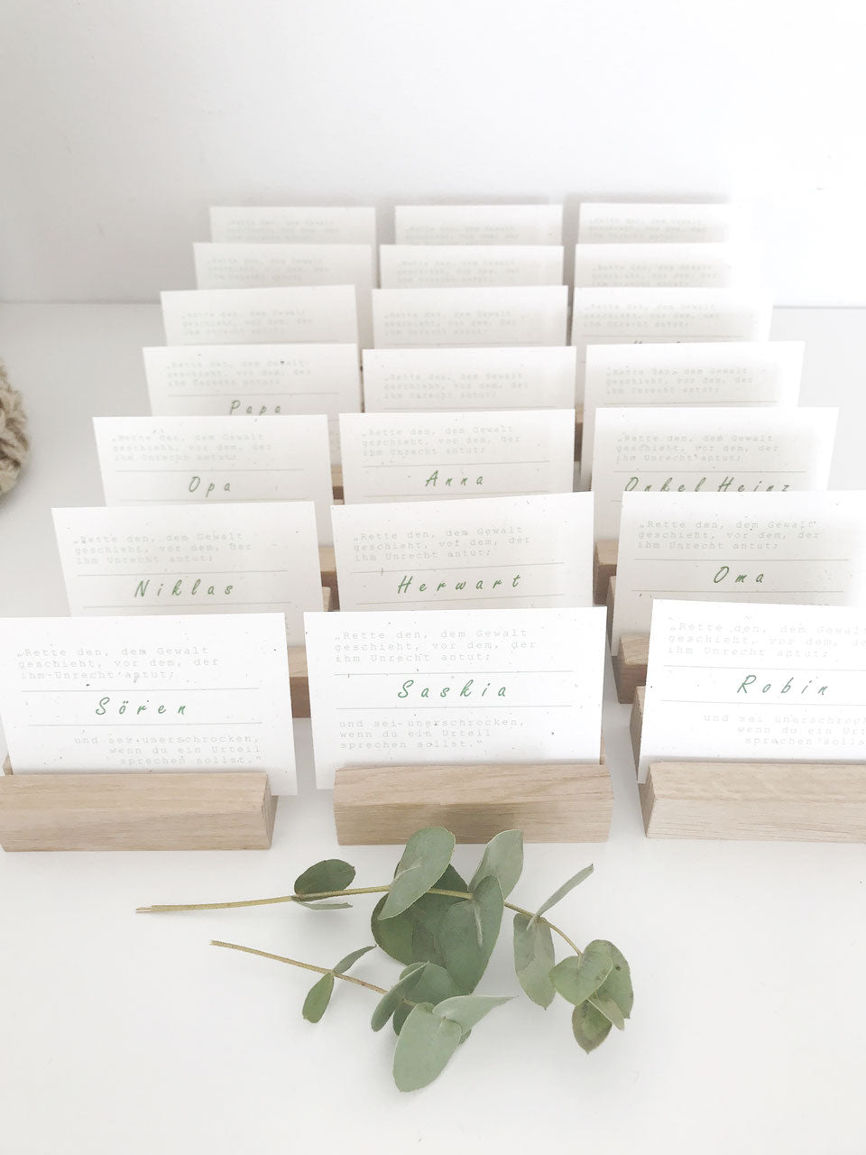 Drei Reihen mit hintereinander stehenden Platzkarten mit Namen in kleinen Kartenhaltern aus Eichenholz. Davor liegt ein Zweig Eukalyptusblätter