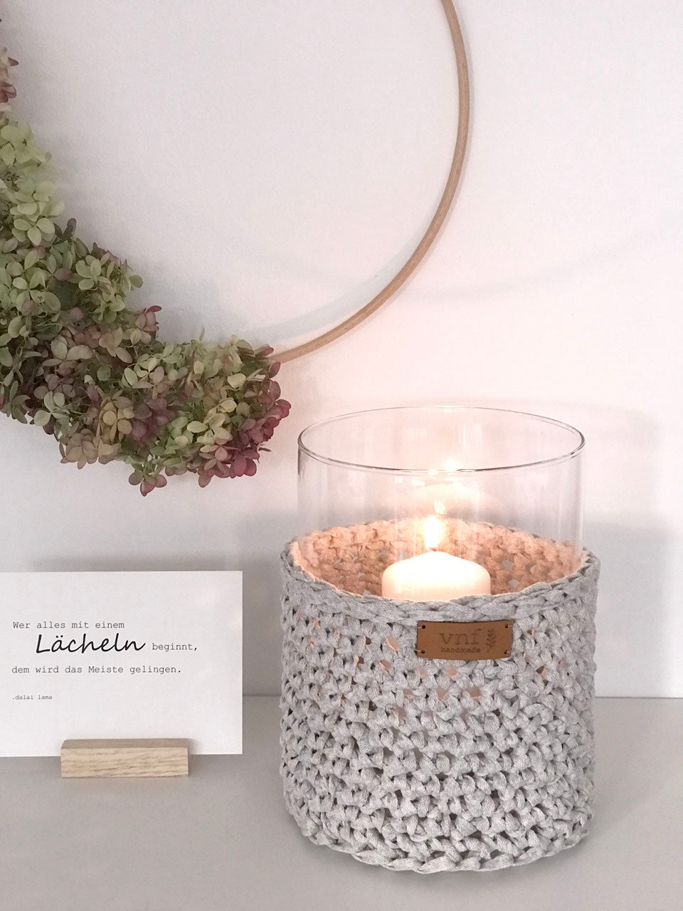 Zylindrische Glasvase mit einem gehäkeltem Überzug in leinen, gefüllt mit Weidenzweigen auf einer Holzbank, daneben gebündelt vier braune Kerzen