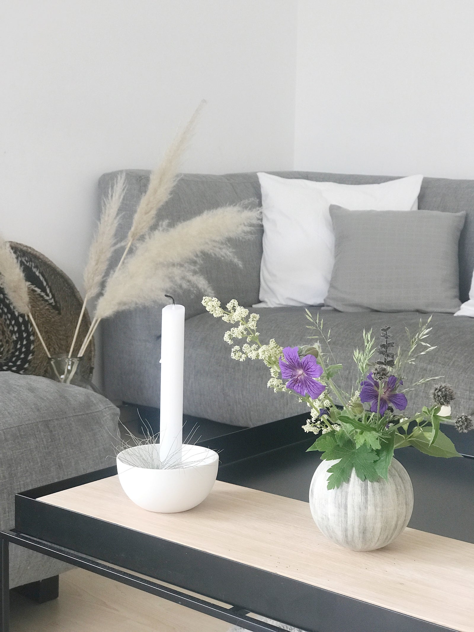Wohninspiration: im Hintergrund ein graues Sofa mit grauen und weißen Kissen, Trockengräsern. Davor ein schwarzer Metalltisch mit einem Holztablett, einem weißen Kerzenhalter, einer Vase mit Gartenblumen. Natürlich, schlichte Einrichtung