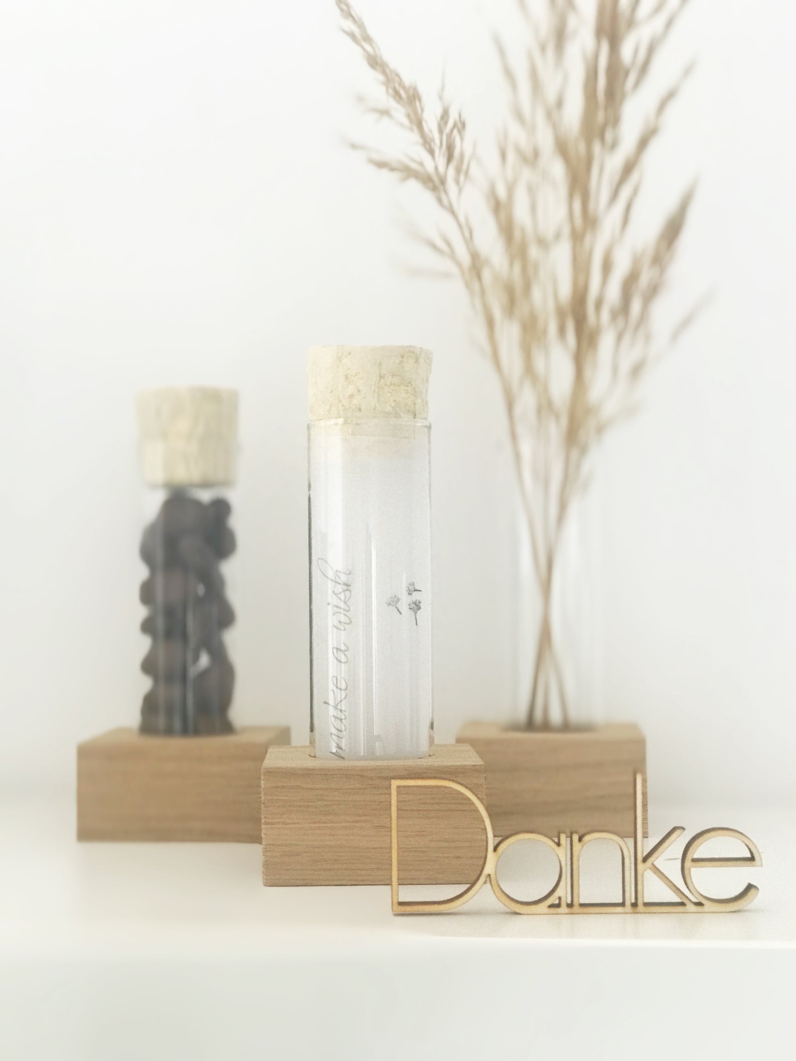 Drei große Reagenzgläser mit Naturkorken in einer Halterung us Eichenholz. Die Gläser sind mit Schokodrops, Trockenblumen und einem Transparentpapier gefüllt. Davor steht ein gelaserter Holz Schriftzug "Danke"